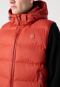 Rød puffervest med hette, har glidelås foran, sidelommer og et rundt logo. Laget av glatt, isolert stoff.