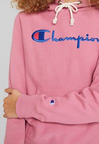 Sweatshirt rose avec une capuche à cordon de serrage et un logo "Champion" brodé en bleu. Comprend une poche avant et un petit logo sur la manche.