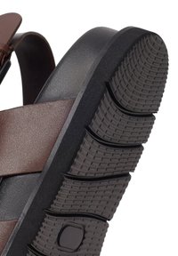 Sandaal met bruine leren bandjes en dikke zwarte rubberen zool met diepe flexgroeven en een getextureerd profiel.