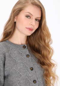 Grau gestrickte Strickjacke mit einem strukturierten Muster, rundem Ausschnitt und dunklen Knöpfen. Weicher Stoff mit dezentem Zopfmuster.