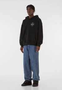 Black hoodie with front pocket, logo graphic on chest, worn with loose blue jeans and black sneakers. Smooth fabric, casual fit.