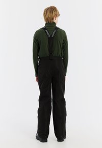 Zwarte sneeuwbroeken met verstelbare bretels, gecombineerd met een donkergroen langemouwen shirt. De broeken hebben een losse pasvorm en een duurzame textuur.