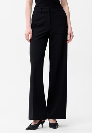 Kobieta ubrana w czarne spodnie z szerokimi nogawkami i czarne szpilki z zaostrzonymi noskami, stojąca na białym tle.