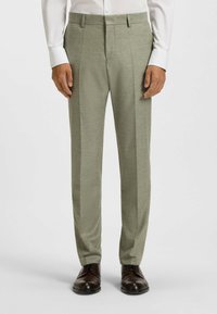 Jasnozielone dopasowane spodnie o teksturowanej tkaninie, z dwoma plisami z przodu i kieszeniami po bokach, w zestawieniu z ciemnobrązowymi butami.