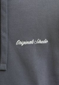 Tejido gris oscuro con una superficie texturizada y acento tejido. Presenta el texto bordado en blanco "Originals Studio".