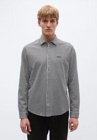 Garām piedurknēm kreklā ar melnām un baltām vertikālām svītrām, kas ir ar smailu apkakli, pogātu priekšpusi un mazu logotipu pa kreisi krūtīs.