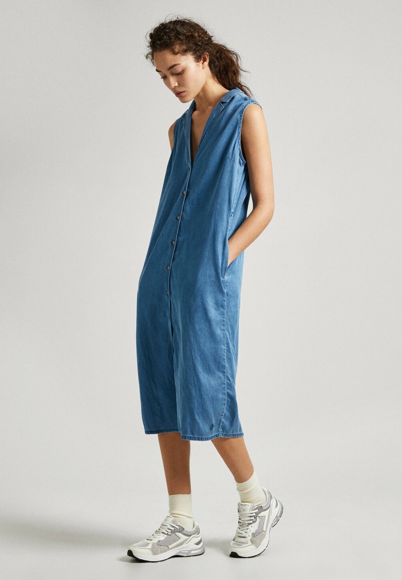 Robe en denim sans manches de couleur bleu clair avec un décolleté en V profond, fermeture à boutons, poches latérales et une coupe ample à mi-mollet. Assortie à des baskets sportives.