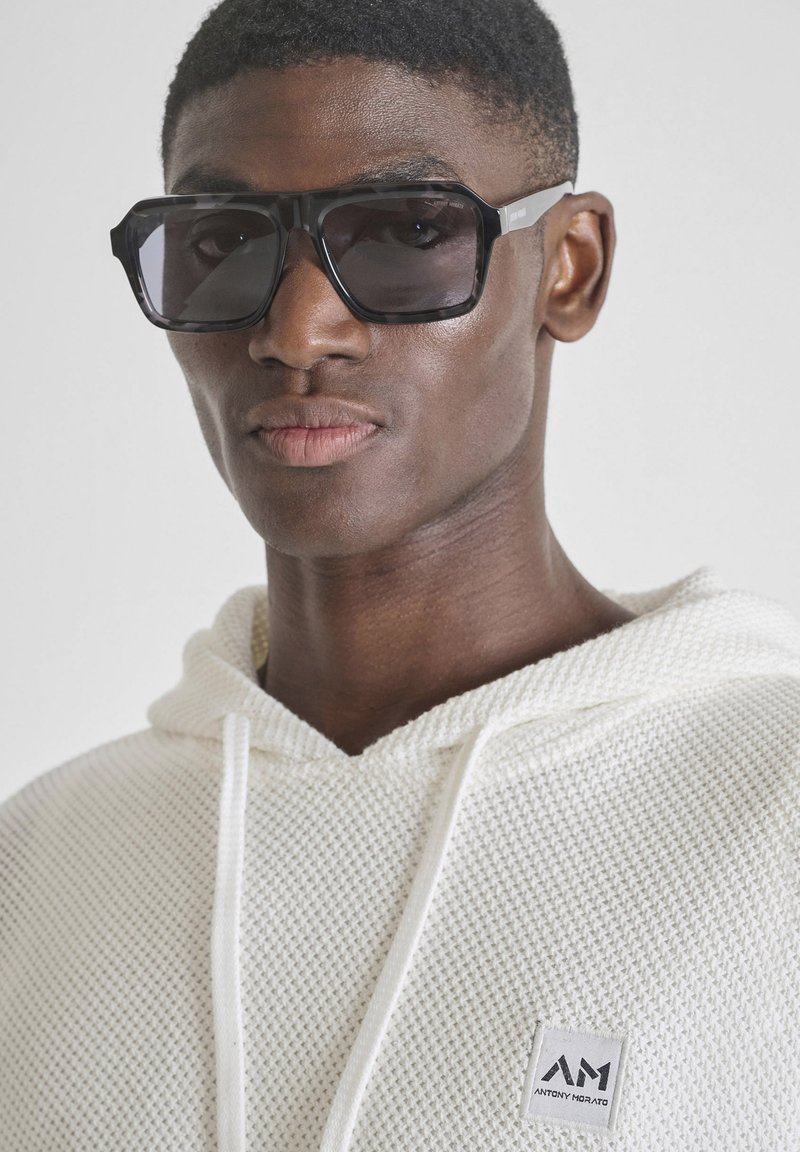 Hombre que lleva unas grandes gafas de sol cuadradas negras y una sudadera blanca con textura con un pequeño logo de Antony Morato en el pecho.