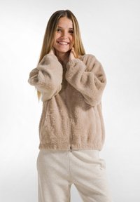 Chaqueta de felpa beige con cremallera y mangas abullonadas, con una textura suave, llevada sobre pantalones brillantes de color claro. Diseño simple y casual.