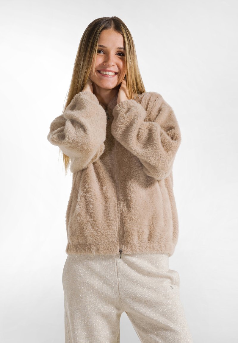 Chaqueta de felpa beige con cremallera y mangas abullonadas, con una textura suave, llevada sobre pantalones brillantes de color claro. Diseño simple y casual.
