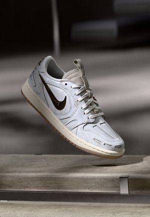 Hvid Nike-sneaker med gyldent swoosh, der svæver over en betonbænk i en blødt oplyst indendørssituation.