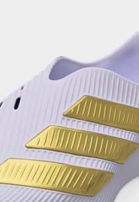 Gros plan sur une chaussure de sport blanche avec une surface texturée et trois bandes parallèles dorées sur le côté.