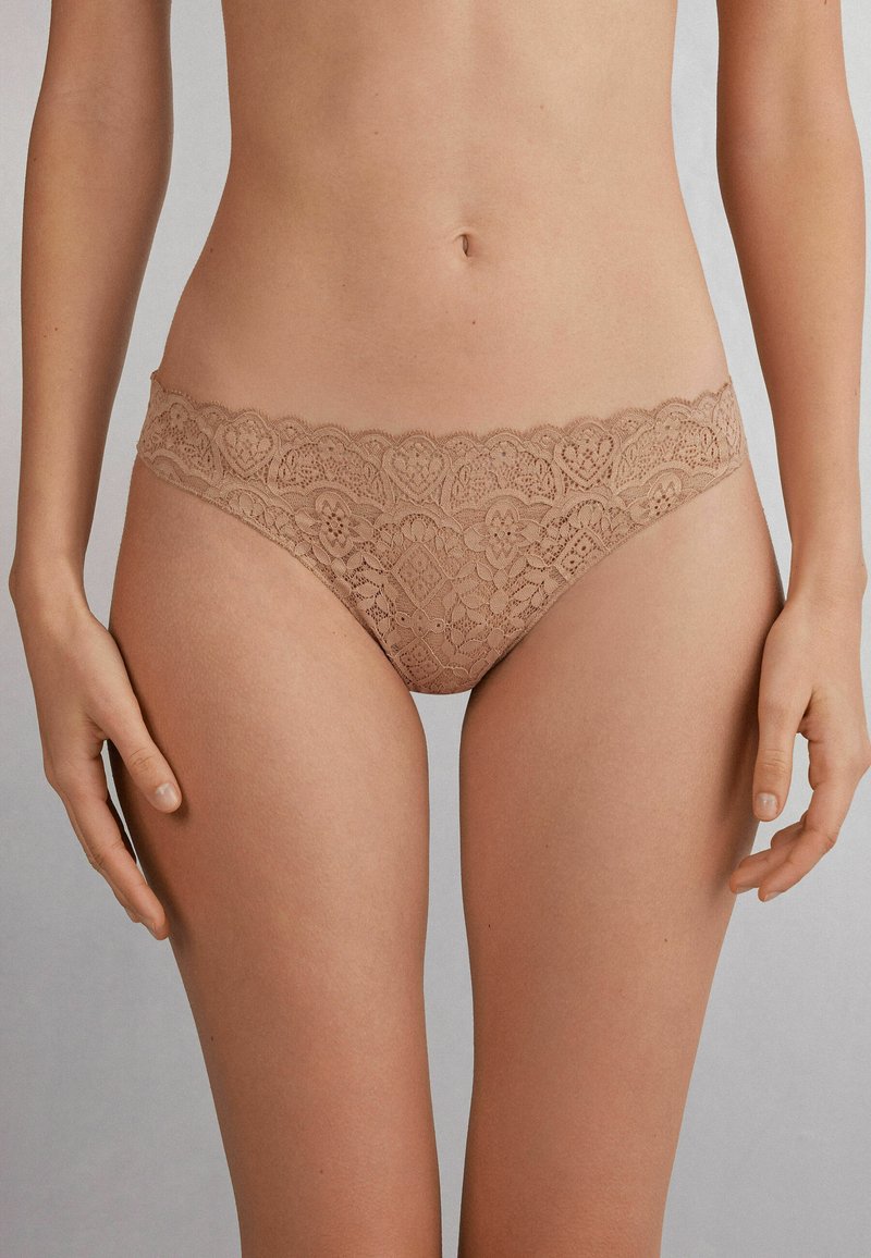 String en dentelle beige avec bords festonnés, présentant des motifs élaborés et une texture douce. Conçu pour un ajustement confortable et une couverture minimale.