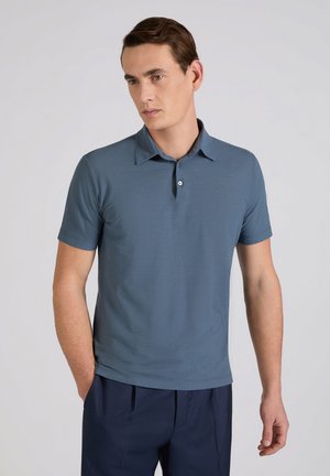 Uomo con capelli castani corti che indossa una polo a maniche corte blu e pantaloni blu scuro, in piedi con una mano in tasca.