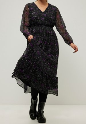 Frau trägt ein schwarzes Midikleid mit lila Blumenmuster, transparente lange Ärmel, hält den Rock, kombiniert mit schwarzen, mit Nieten besetzten Stiefeletten.