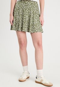 Jupe florale verte avec des motifs blancs, design évasé, tissu léger, associée à des baskets blanches et des chaussettes.