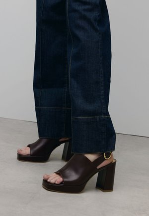 Sandales en cuir marron à plateforme avec orteils ouverts, talons imposants et un accent métallique circulaire, portées avec un pantalon en denim foncé à pattes d'éléphant.