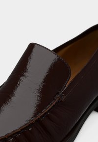 Σκούρο καφέ γυαλιστερό δερμάτινο μοκασίνι με λεπτομέρειες ραφής και χαμηλό τακούνι σε λευκό φόντο.