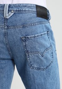 Light blue denim jeans featuring a v-shaped back pocket, distinct stitching, and a black leather patch on the waistband with brand details.