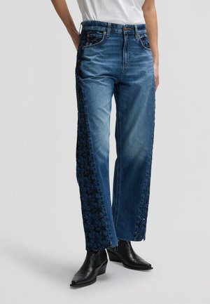 Siniset leveälahkeiset farkut, joissa mustat kukkakuvioiset pitsipaneelit sivuilla, yhdistettynä mustiin nilkkureihin ja valkoiseen yläosaan.