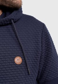 Navyblauwe gestructureerde hoodie met een capuchon, contrasterende trekkoorden en een ronde leren accent met reliëf tekst op de borst.