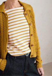 Person som bär en senapsgul stickad cardigan över en vit och gulrandig skjorta som är instoppad i mörka jeans med händerna i fickorna.