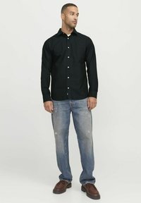 Schwarzes Hemd mit langen Ärmeln, leichtes Material, kombiniert mit hellblauen, verwaschenen Jeans und dunkelbraunen Schuhen. Einfache Taschenverzierung.