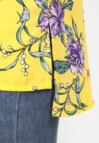 Blusa gialla con stampe floreali viola, foglie e dettagli bianchi. Tessuto morbido con una forma leggermente svasata e spacchi laterali.