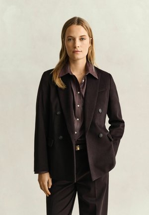 Femme aux longs cheveux blonds portant un blazer croisé sombre, un pantalon assorti et une chemise rayée sombre boutonnée, se tenant devant un fond clair.