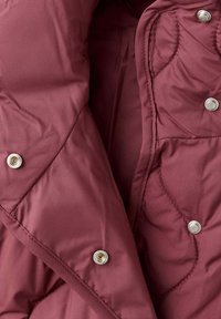 Gros plan sur un tissu matelassé bordeaux avec des boutons-pression argentés sur une veste ou un manteau matelassé.