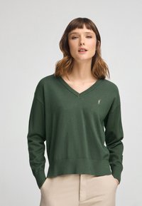 Pull en V vert foncé avec des manches longues, bords-côtes aux poignets et à l'ourlet, et un petit logo brodé sur la poitrine. Texture lisse et douce.