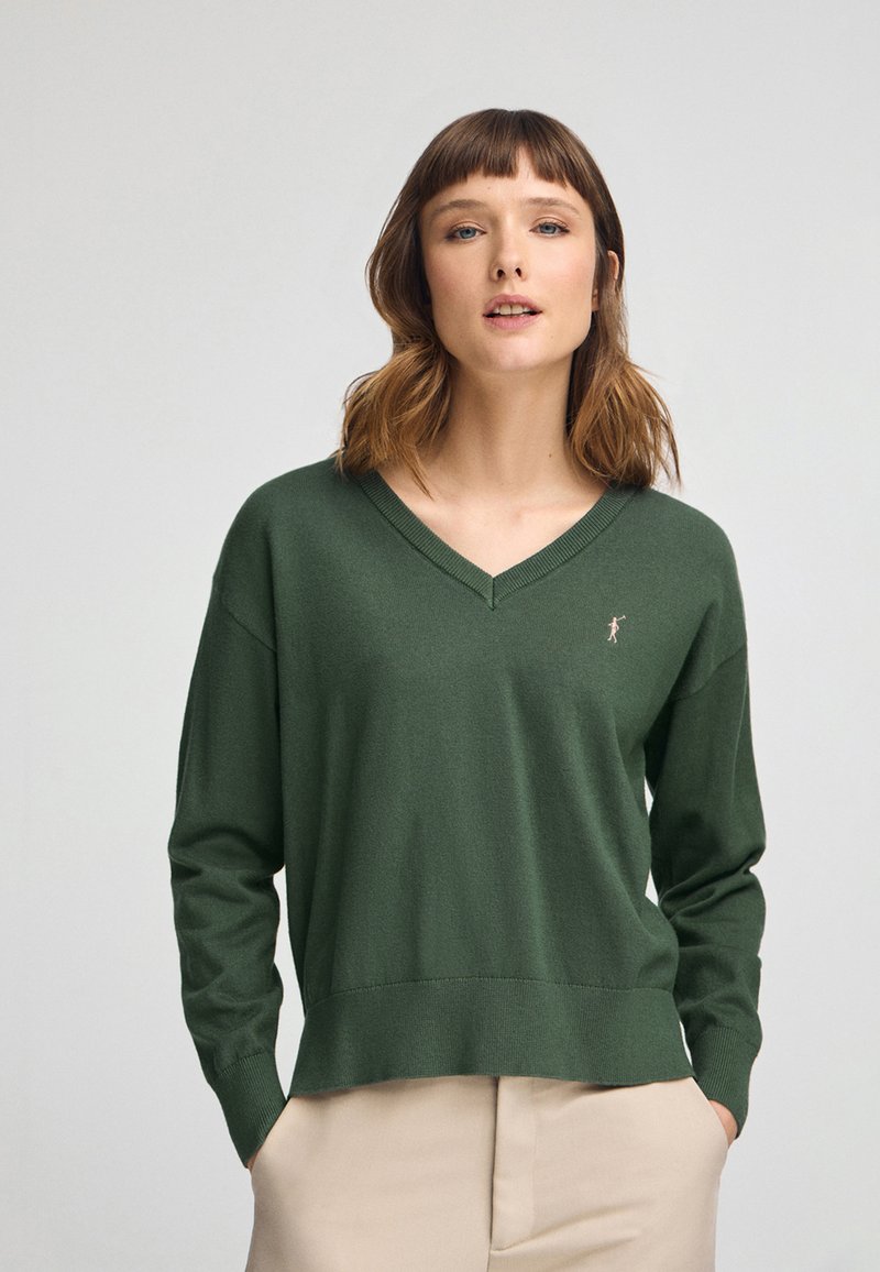 Pull en V vert foncé avec des manches longues, bords-côtes aux poignets et à l'ourlet, et un petit logo brodé sur la poitrine. Texture lisse et douce.