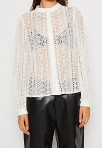 Vit spetsblus med långa ärmar, mandarin-krage, knappstängning och genomskinlig design, kombinerad med svarta läderbyxor.