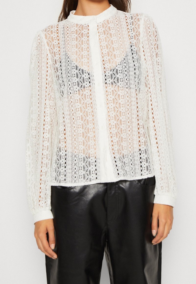 Vit spetsblus med långa ärmar, mandarin-krage, knappstängning och genomskinlig design, kombinerad med svarta läderbyxor.