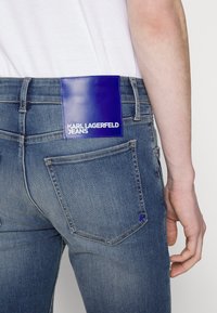 Obepaslé modré džíny s výraznou fialovou nášivkou „KARL LAGERFELD JEANS“ na zadní kapse, které nosí osoba ve белém tričku.