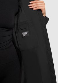 Zwarte gewatteerde jas met een rits aan de voorkant, voorzien van twee voortassen en een stoffen label aan de linkerkant, gedragen over een zwarte top.