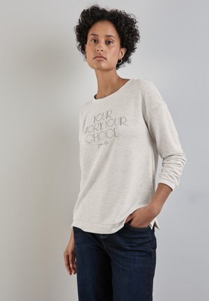 Hellgrau sweatshirt mit langen Ärmeln und einem Rundhalsausschnitt, versehen mit auffälligem schwarzem Text. Kombiniert mit dunkelblauen Jeans, die legere Kleidung demonstrieren.