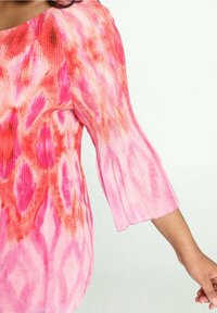 Donna che indossa una blusa a tie-dye rosa e rossa con maniche a tre quarti plissettate e svasate, su uno sfondo chiaro e semplice.