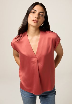 Blusa color salmone a maniche corte con scollo a V profondo, realizzata in tessuto morbido, caratterizzata da una vestibilità rilassata e orlo curvo. Abbinata a jeans blu.