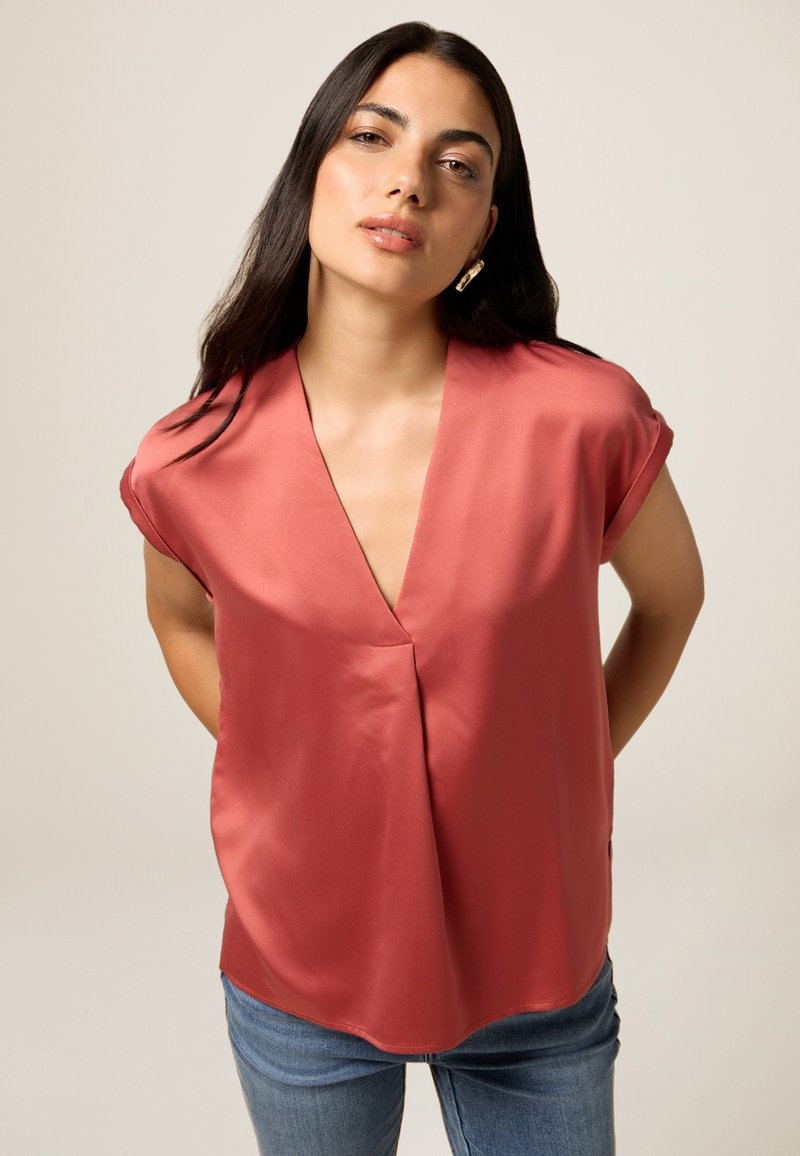 Blusa color salmone a maniche corte con scollo a V profondo, realizzata in tessuto morbido, caratterizzata da una vestibilità rilassata e orlo curvo. Abbinata a jeans blu.