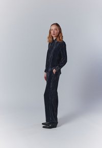 Tengerészes plisszírozott ing és nadrág szett texturált anyagból, hosszú ujjakkal, gombos záródással és oldalsó zsebekkel, fekete magassarkú csizmával párosítva.