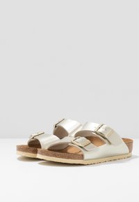 Metalliskt gyllene sandaler med två justerbara remmar och silvriga spännen. Korkfotbädd och ljusbrun gummiyttersula. Öppen design för bra luftgenomströmning.