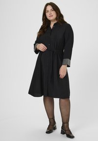 Vrouw met lang bruin haar in een zwarte jurk tot de knie met knopen aan de voorkant, gebloemde manchetten, transparante zwarte panty en zwarte hakken die staat.