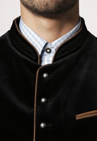 Schwarze Samtjacke mit einem strukturierten Kragen, silbernen Knöpfen und braunen Akzenten; darunter trägt sie ein hellblaues und weißes Karoshirt.