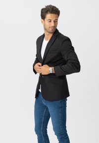 Sort blazer med klapflip, enkelt knaplukning, to forlommer, parret med en hvid t-shirt og blå jeans. Glat tekstur.