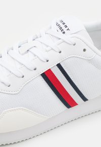 Białe sneakersy z chropowatą tkaniną na cholewce, z czerwonymi i granatowymi paskami oraz gładką gumową podeszwą. Widoczne sznurowadła oraz logo na języku.