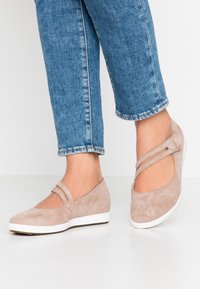 Zapatos planos de ante beige con lados recortados y detalles de dos correas, que cuentan con una suela de goma blanca y una textura suave. Pantalones de mezclilla cortos visibles.