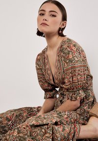 Robe à motifs floraux dans un vert terreux avec des manches courtes bouffantes, un décolleté en V profond et des accents froncés. Sandales compensées en liège, texture douce.