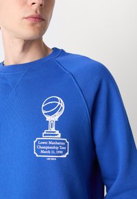 Blauwe sweatshirt met een ronde hals en raglanmouwen, gekenmerkt door een witte afbeelding van een trofee en de tekst: "Lower Manhattan Championship Tour 11 maart 1990."