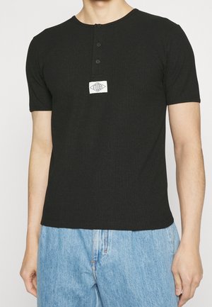 Homme portant un t-shirt Henley noir à manches courtes côtelé avec trois boutons et un logo blanc centré, associé à un jean bleu clair.